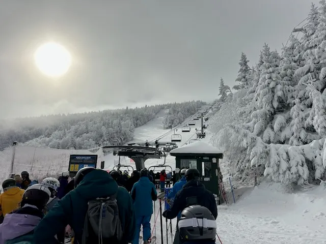Skiers lined up for a chairlift beneath snow-covered evergreens in early November