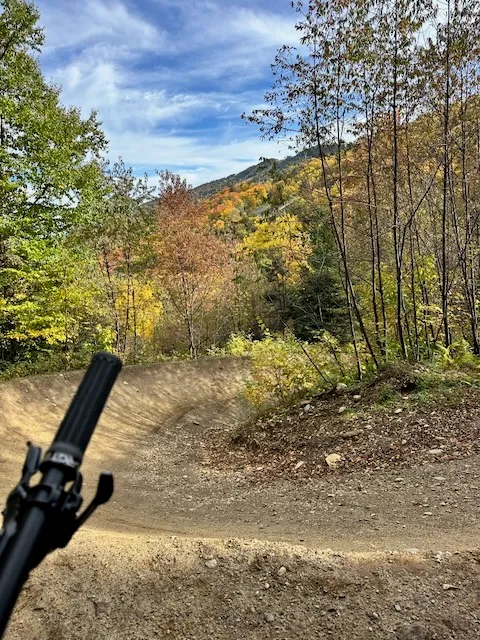 Bike park berm turn with a handlebar in the foreground and fall foliage on the mountainside beyond