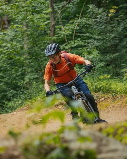 Mountain biker in an orange shirt and helmet leaning into a turn on a dirt trail through green woods