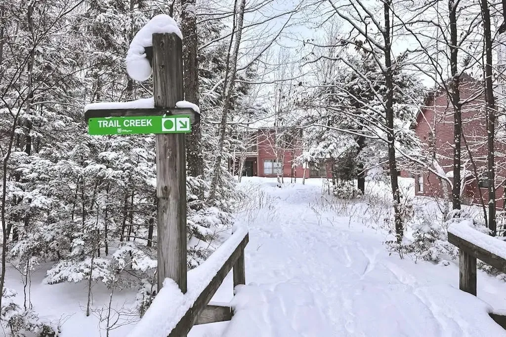 Snow-covered path with a Trail Creek trail sign leading toward the condo buildings