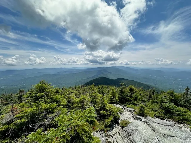 Summit view over spruce and rock toward Mount Mansfield on the horizon under a partly cloudy sky