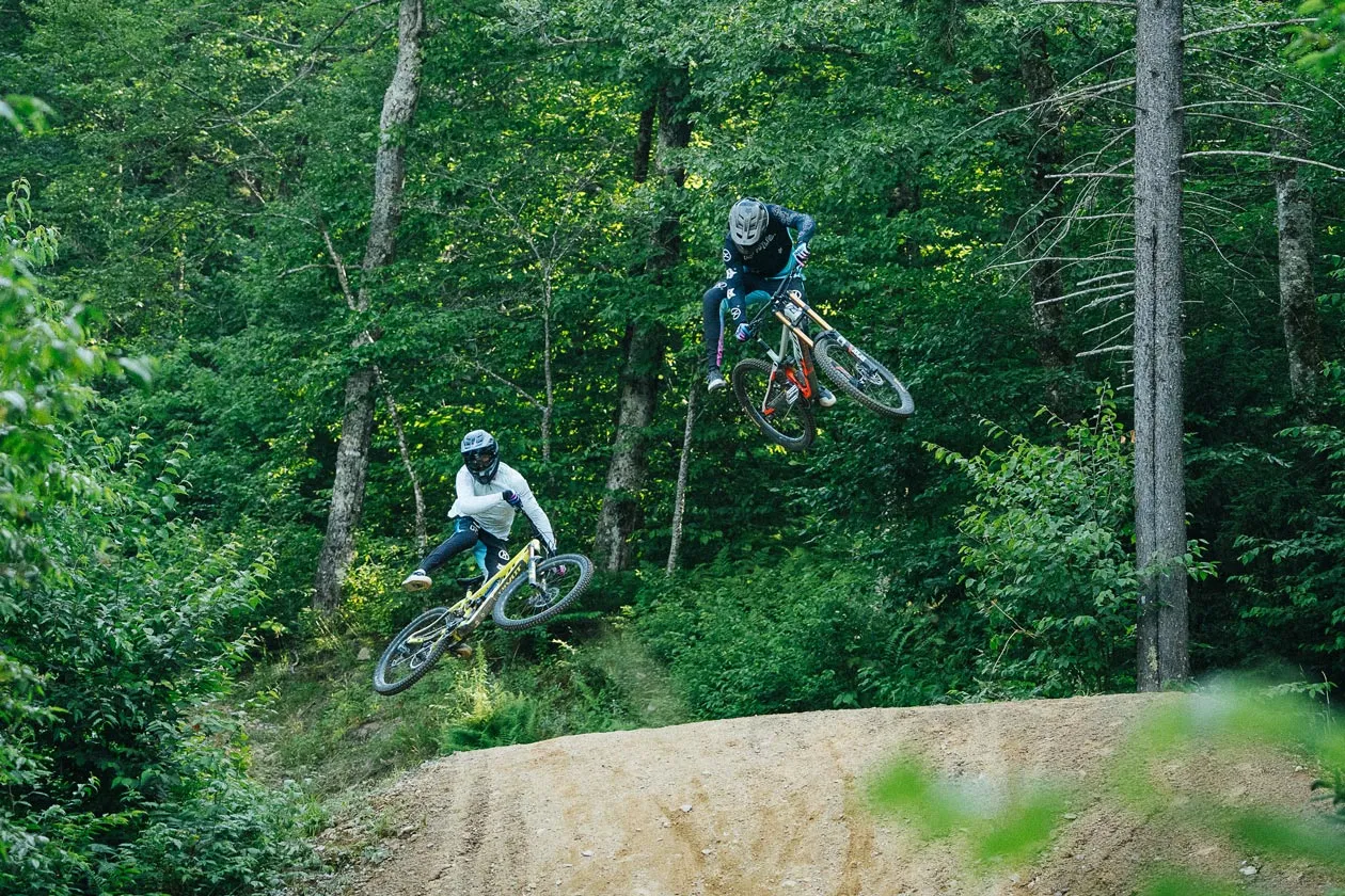 Two mountain bikers airborne over a dirt jump in a green forest