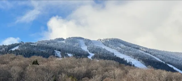 Snow-dusted ski trails on a forested mountainside under a partly cloudy sky in November